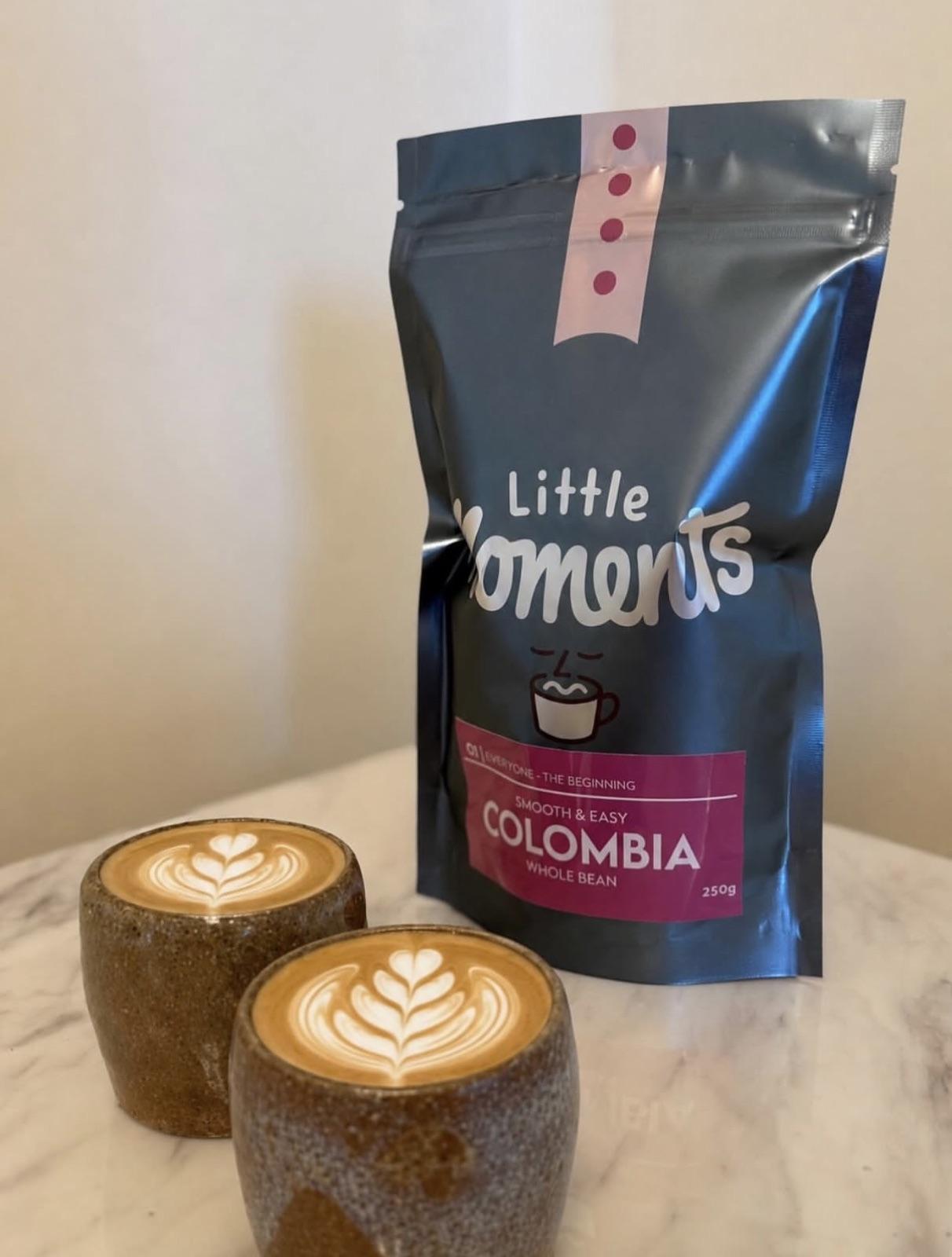 A Little Moments bag beside two flat whites with latte art on a marble table