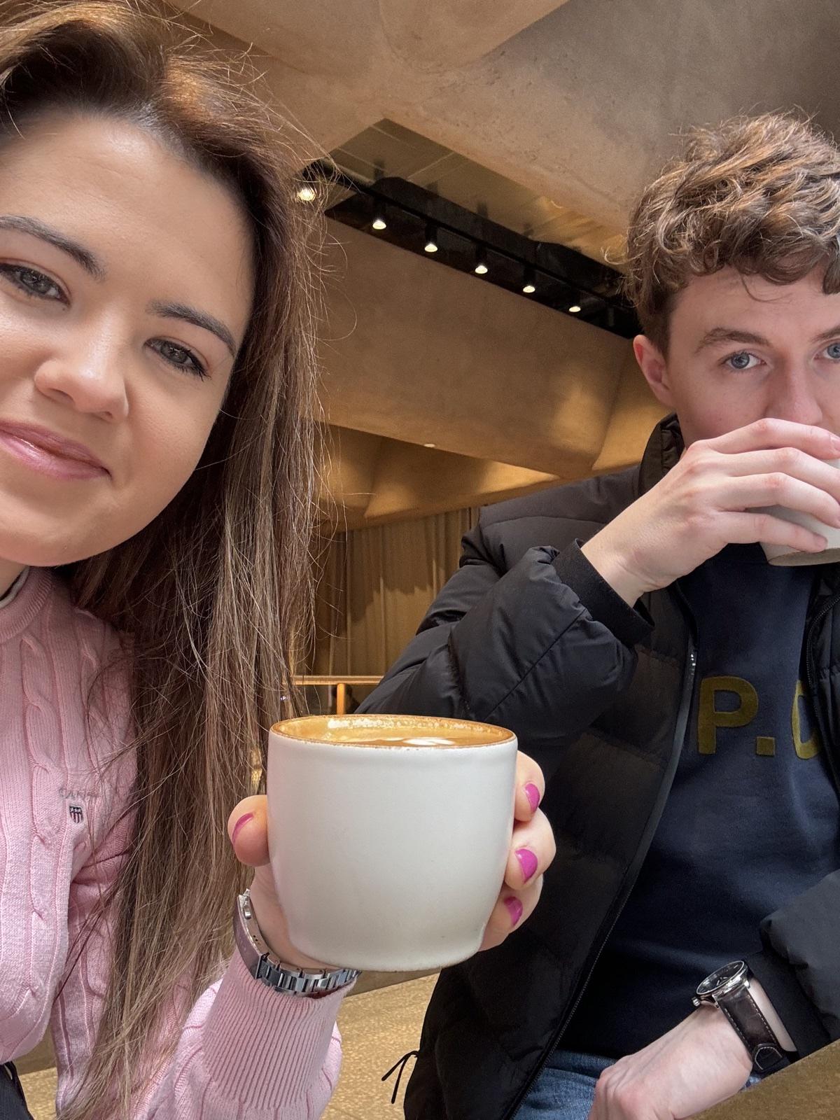 Erin holding up a cup of coffee at a café table, with company beside her